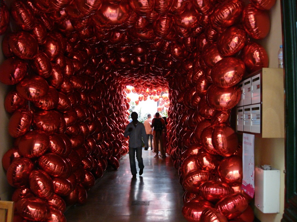 Red balloons lining the walls and ceilings of the corridor leading to the interior courtyard where the balloons are suspended in the air.