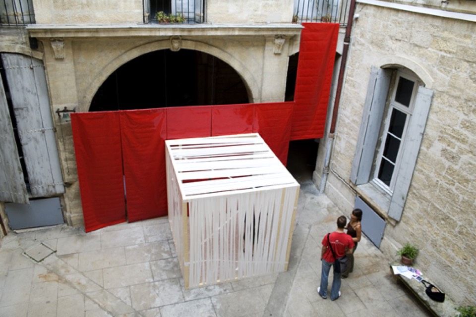 Red tarps suspended from courtyard openings flanking a wooden box wrapped in white tape.