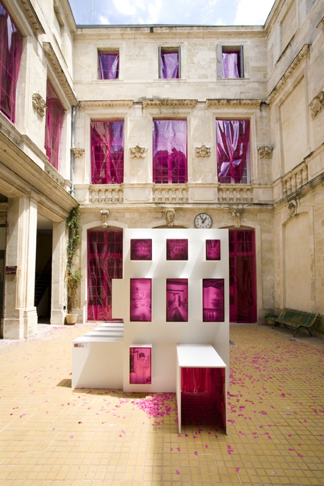White box structure with openings covered in pink cellophane in the center of the courtyard. Courtyard thresholds also covered with pink cellophane.