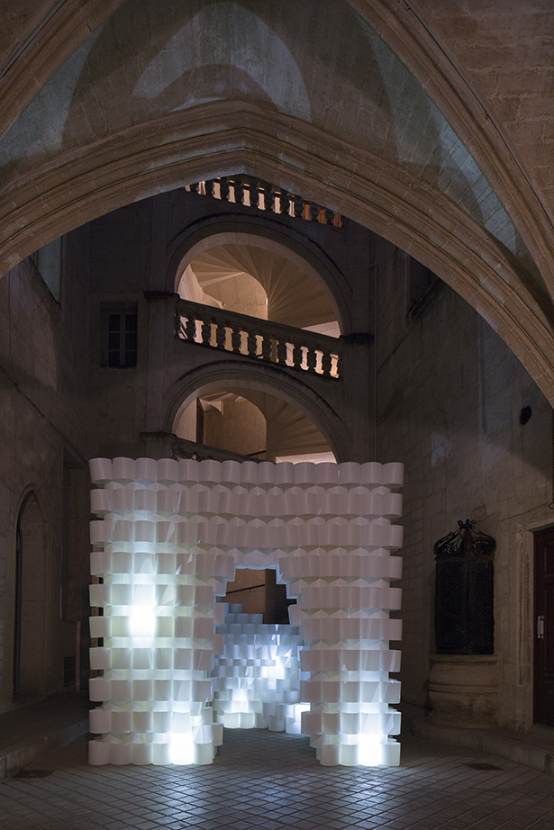 An illuminated wall of stacked white paper cylinders stands within a dark stone courtyard